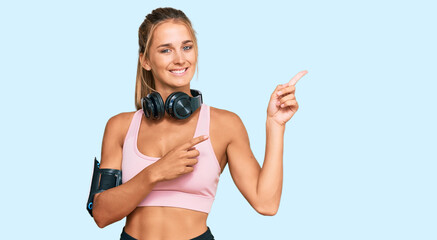 Wall Mural - Young blonde woman wearing gym clothes and using headphones smiling and looking at the camera pointing with two hands and fingers to the side.