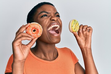 Sticker - Young african american woman holding tasty colorful doughnuts on eyes angry and mad screaming frustrated and furious, shouting with anger looking up.