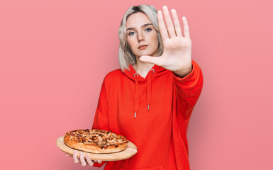 Poster - Young blonde girl holding italian pizza with open hand doing stop sign with serious and confident expression, defense gesture