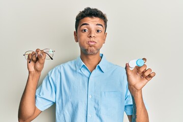 Sticker - Young handsome african american man holding glasses and contact lenses puffing cheeks with funny face. mouth inflated with air, catching air.