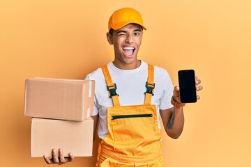 Poster - Young handsome african american man holding delivery package and showing smartphone screen winking looking at the camera with sexy expression, cheerful and happy face.