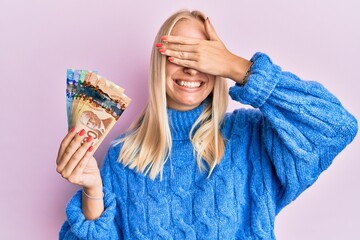 Sticker - Young blonde girl holding canadian dollars smiling and laughing with hand on face covering eyes for surprise. blind concept.