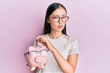 Sticker - Young chinese woman holding piggy bank with glasses clueless and confused expression. doubt concept.