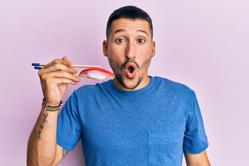 Poster - Handsome man with tattoos eating tuna sushi using chopsticks scared and amazed with open mouth for surprise, disbelief face