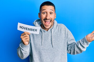 Canvas Print - Handsome man with tattoos holding november word on paper celebrating achievement with happy smile and winner expression with raised hand