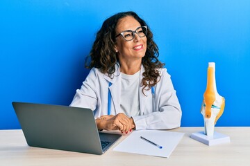 Poster - Beautiful middle age woman doctor at orthopedic clinic looking away to side with smile on face, natural expression. laughing confident.