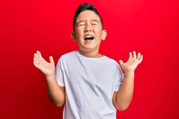 Wall Mural - Little boy hispanic kid wearing casual white tshirt celebrating mad and crazy for success with arms raised and closed eyes screaming excited. winner concept