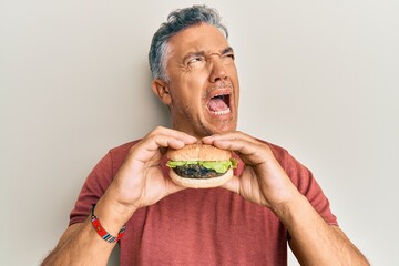 Wall Mural - Handsome middle age mature man eating a tasty classic burger angry and mad screaming frustrated and furious, shouting with anger. rage and aggressive concept.