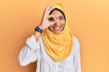 Canvas Print - Young brunette arab woman wearing traditional islamic hijab scarf doing ok gesture with hand smiling, eye looking through fingers with happy face.