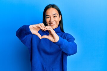 Wall Mural - Young asian woman wearing casual winter sweater smiling in love doing heart symbol shape with hands. romantic concept.
