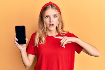 Wall Mural - Beautiful young caucasian girl holding smartphone showing screen afraid and shocked with surprise and amazed expression, fear and excited face.