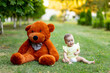 © Any Grant - Cute little girl sitting on green grass with big Teddy bear in yellow summer dress in summer, a place for text
