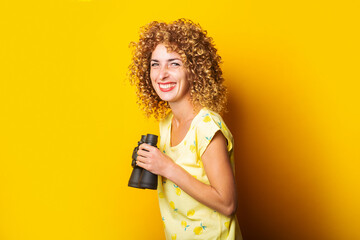 Wall Mural - cheerful smiling curly girl holding binoculars in hands on a yellow background.