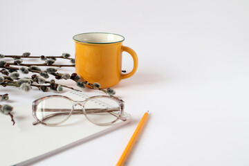 A yellow cup with hot tea, willow branches, a notebook, a pencil, glasses for vision on a white background, a place for text - the concept of an interesting pastime of spring days for the elderly