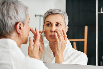 Mature woman applying moisturizer in bathroom. Senior female in bathrobe doing skincare routine.