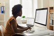 © Studio Romantic - Employee using office computer. African-American woman sitting at desk, looking at desktop PC screen, doing research, studying data reports, working with business documents and online spreadsheets