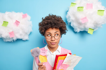 Wall Mural - Photo of pensive Afro American female student holds papers folders and pen makes notes during lecture at university learns material for exam poses against clouds with reminding stickers around