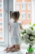 © alexkich - A little girl is sitting on the windowsill. A bouquet of flowers in a vase by the window and a girl sniffing flowers. A little princess in a white dress with a bouquet of white flowers by the window.