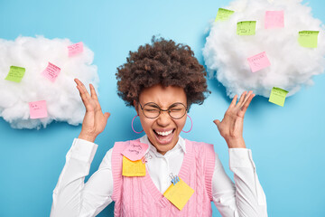 Busy curly Afro American office worker has many tasks at same time exclaims in panic raises hands worries about deadline poses indoor against sticky notes to remember all necessary information