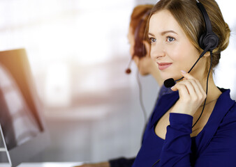 Two busineswomen have conversations with the clients by headsets, while sitting at the desk in a sunny modern office. Diverse people group in a call center. Telemarketing and customer service