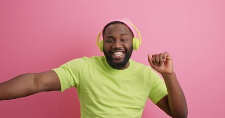 Canvas Print - Bearded cheerful energetic man in headphones has dark skin dances carefree feels very happy raises arms dressed in casual t shirt expresses positive emotions isolated over pink background. Music fan