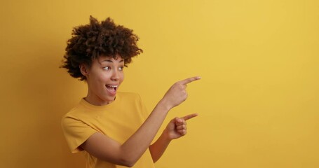 Wall Mural - Positive curly haired woman smiles positively recommends using blank space indicates at upper right corner dressed in casual t shirt isolated over vivid yellow background shows direction or way
