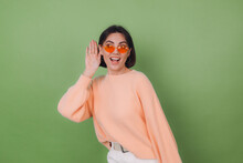 Young Stylish Woman In Casual Peach Sweater And Orange Glasses Isolated On Green Olive Background Curious Try To Hear What You Saying With Hand By Ear Copy Space