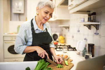 Poster - Attractive gray haired female pensioner in apron standing at kitchen table holding knife, cutting cucumber on board while making new vegetable meal, watching online video recipe on laptop