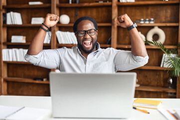 Excited lucky African American student or office manager sitting with laptop at the desk, raised fists up celebrating success, screaming yes, passed test with high score, got promoted, won an award