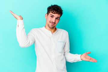 Wall Mural - Young caucasian man isolated on blue background makes scale with arms, feels happy and confident.