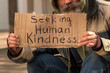 Senior man homeless beggar with a gray beard in a shabby clothes with a carboard sign sitting outdoors in city and asking for money donation. Sign on paperboard Seeking Human Kindness