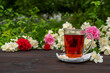 Glass cup with tea on a saucer with two pieces of sugar among flowering bushes of roses and jasmine. Outdoor, picnic, brunch. Green leaves background in blur.