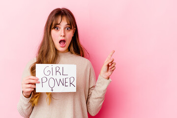 Wall Mural - Young caucasian woman holding a girl power placard isolated on pink background pointing to the side