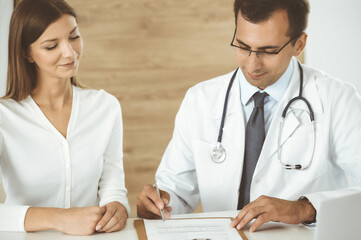 Doctor and patient discussing medical exam results while sitting at the desk in clinic. Male physician using clipboard for filling up medication history record of young woman. Data in medicine