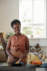 Canvas Print - Young smiling woman of African ethnicity looking at you while standing by electric stove