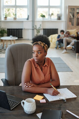 Canvas Print - Pensive young African businesswoman in brown casual blouse making notes in document by table