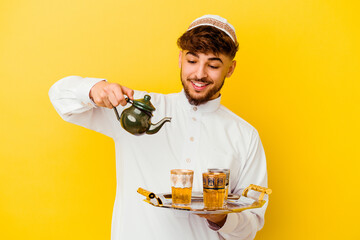 Young Moroccan man wearing the typical arabic costume drinking tea isolated on yellow background