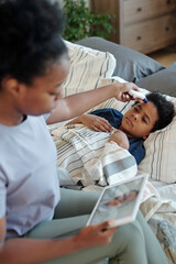 Canvas Print - Young African female measuring body temperature of her sick little son lying in bed under blanket