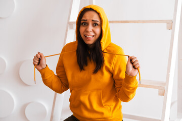 Wall Mural - Portrait of young woman posing on white background. Pretty brunette in yellow hoodie near stepladder on background of white wall.