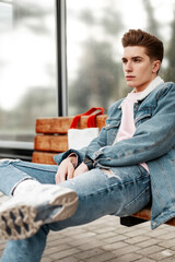 Wall Mural - Trendy young man in youth stylish denim jacket in casual vintage jeans with fabric shopper sit on wooden bench at stop of public building in city. Good-looking guy in fashionable clothes on street.