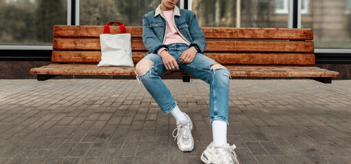 Wall Mural - Trendy young man in youth stylish denim jacket in casual vintage jeans with fabric shopper sit on wooden bench at stop of public building in city. Good-looking guy in fashionable clothes on street.