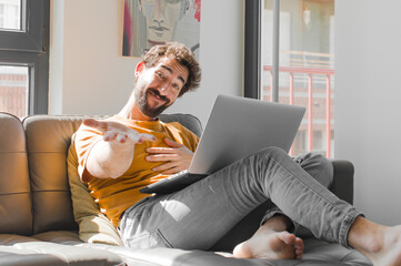 Wall Mural - young bearded man smiling happily with friendly, confident, positive look, offering and showing an object or concept with a laptop on a couch laptop concept