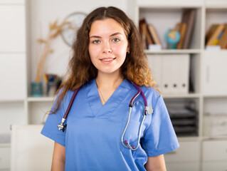 Wall Mural - Friendly female doctor in a blue shirt with a stethoscope standing in medical office in a clinic