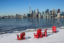 Red Muskoka Chairs With Toronto Skyline And Frozen Lake Ontario In Winter