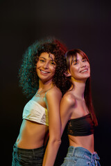 Wall Mural - Two cheerful young brunette female models with professional art makeup smiling, posing together in neon light isolated over black background