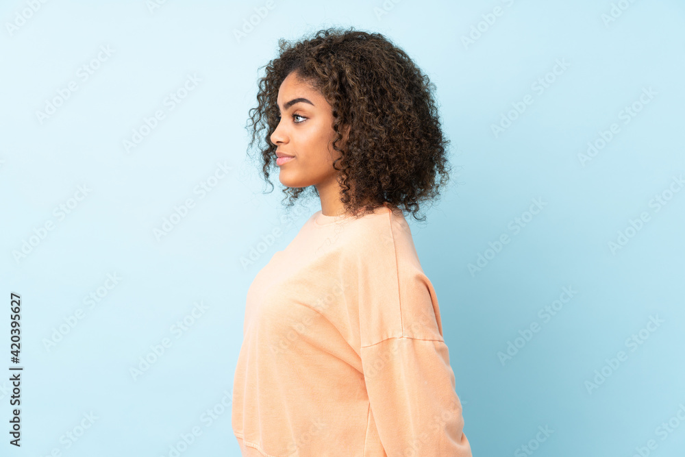 Young African American woman isolated on blue background in back position and looking back - obrazy, fototapety, plakaty 