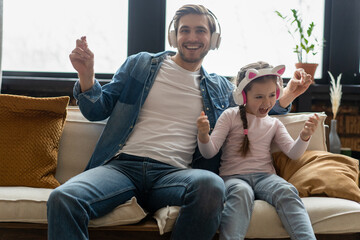 Wall Mural - Cute little girl and her handsome father in headphones are listening to music, smiling, sitting on couch at home.