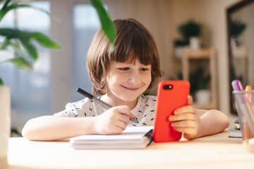 Wall Mural - Back to school. Distance learning online education. Caucasian smile kid boy studying at home with mobile smart phone and doing school homework. Thinking child siting at table with cell phone