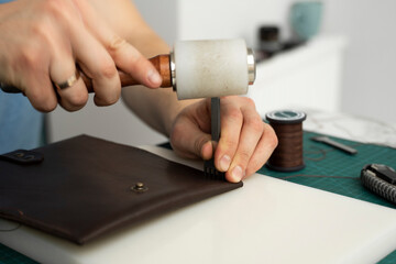 Mens hand holding a leather carving hammer and steel chisel hole punch and makes a leather wallet in his workshop. Working process with a brown natural leather. Craftsman holding a crafting tools.