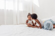 © farmuty - Brother and sister African Americans play together on white bed in loft interior. Siblings having fun among the blue pillows in the morning. Boy kissing little girl on the cheek, caring for a neighbor